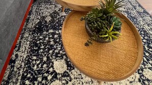 En moderne stue med en grå sofa, to runde træ sofaborde med plantepotte, en sort fjernbetjening og et mønstret tæppe.