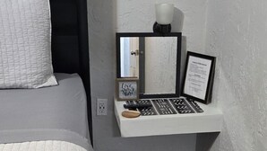 Un dormitorio con cama, una mesita de noche con lámpara, un espejo y un objeto enmarcado en la pared.