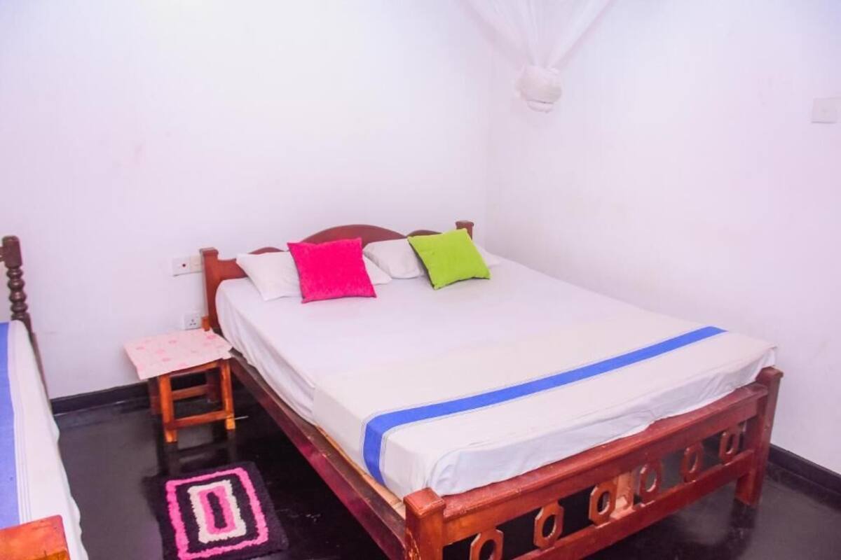 A single bed with a wooden frame, white bedding, and colorful pillows. A small wooden stool is beside the bed, and a mosquito net hangs on the wall.