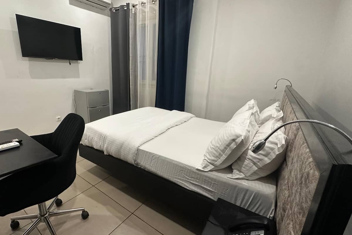 Una camera piccola e pulita con un letto, una scrivania, una sedia, una televisione e una finestra con tende.