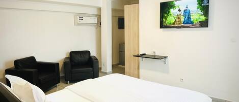 Une chambre d’hôtel avec un lit, deux chaises noires, un téléviseur fixé au mur et une petite étagère.