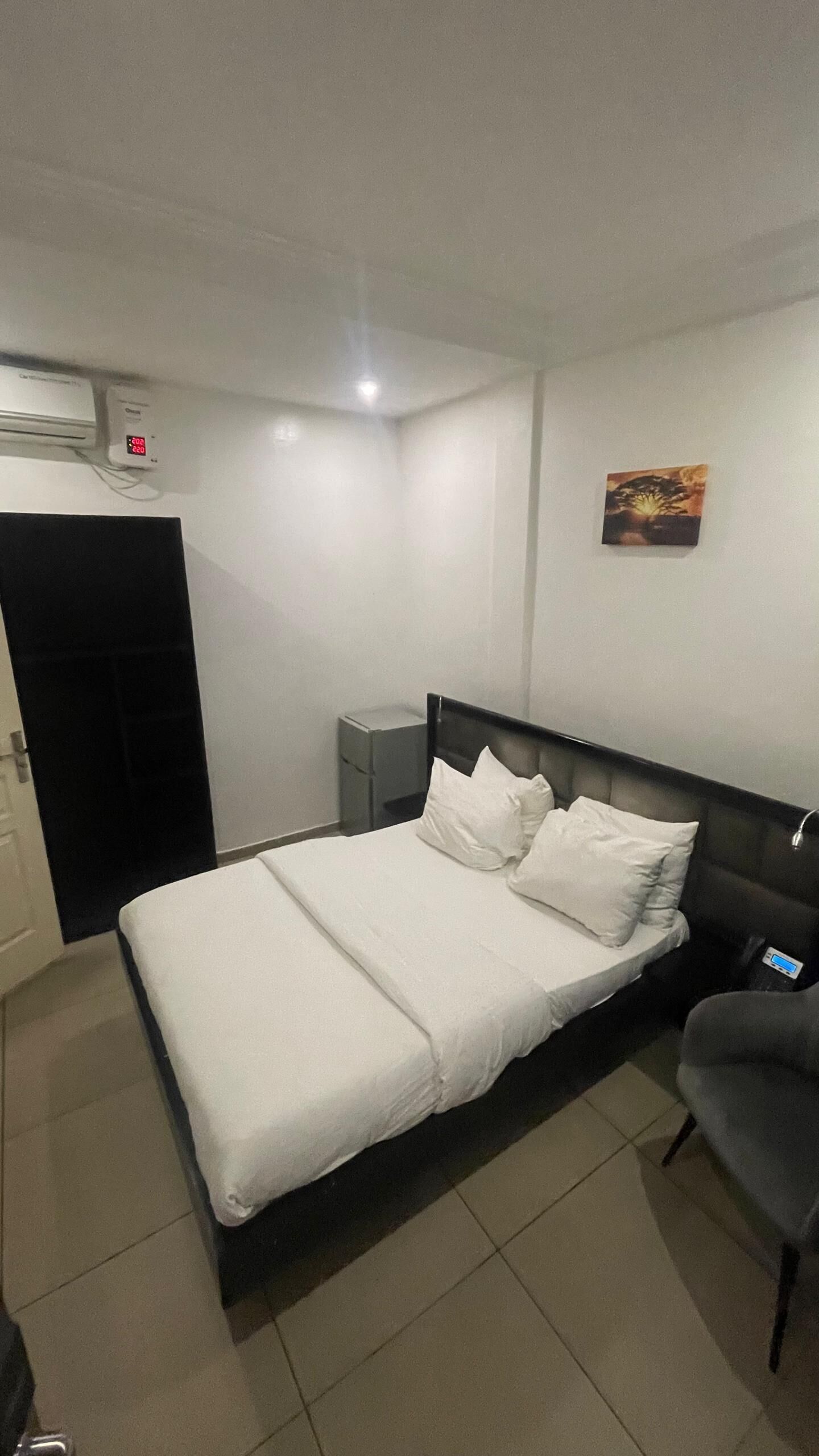 A hotel room with a bed, a chair, a nightstand, a wall-mounted air conditioner, and a small painting.