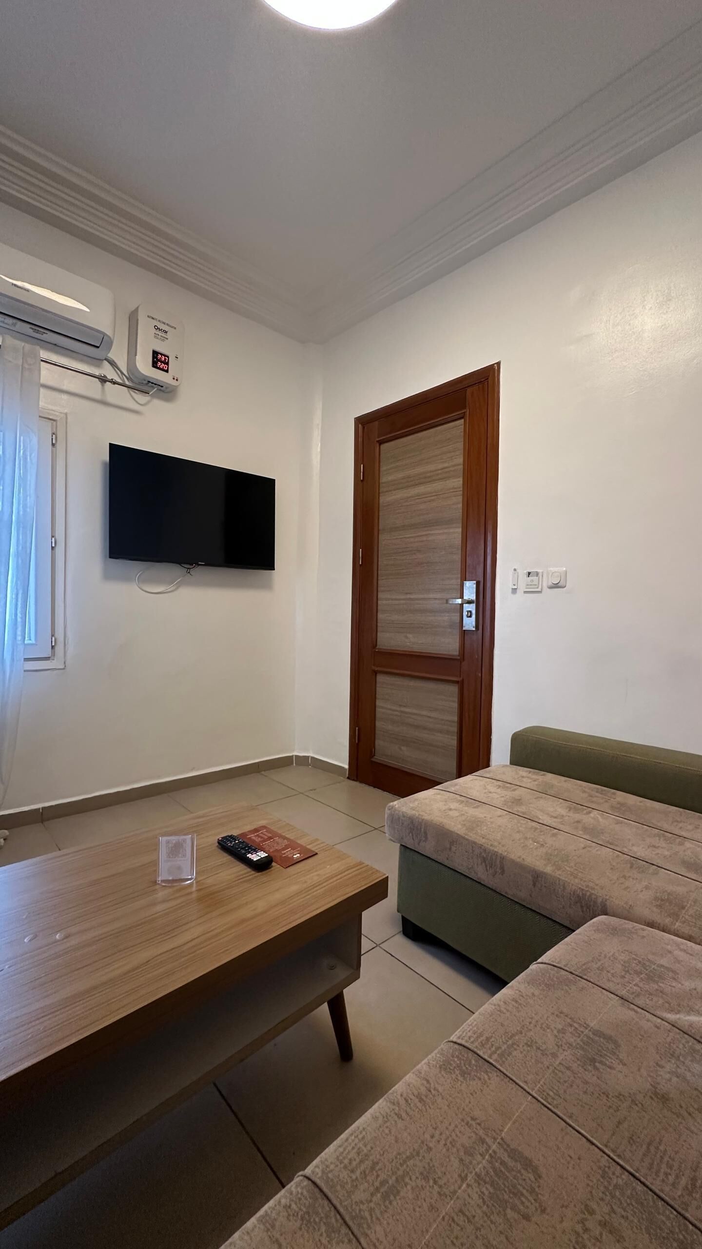 A room with a wooden table, a green sofa, a TV mounted on the wall, and a wooden door.
