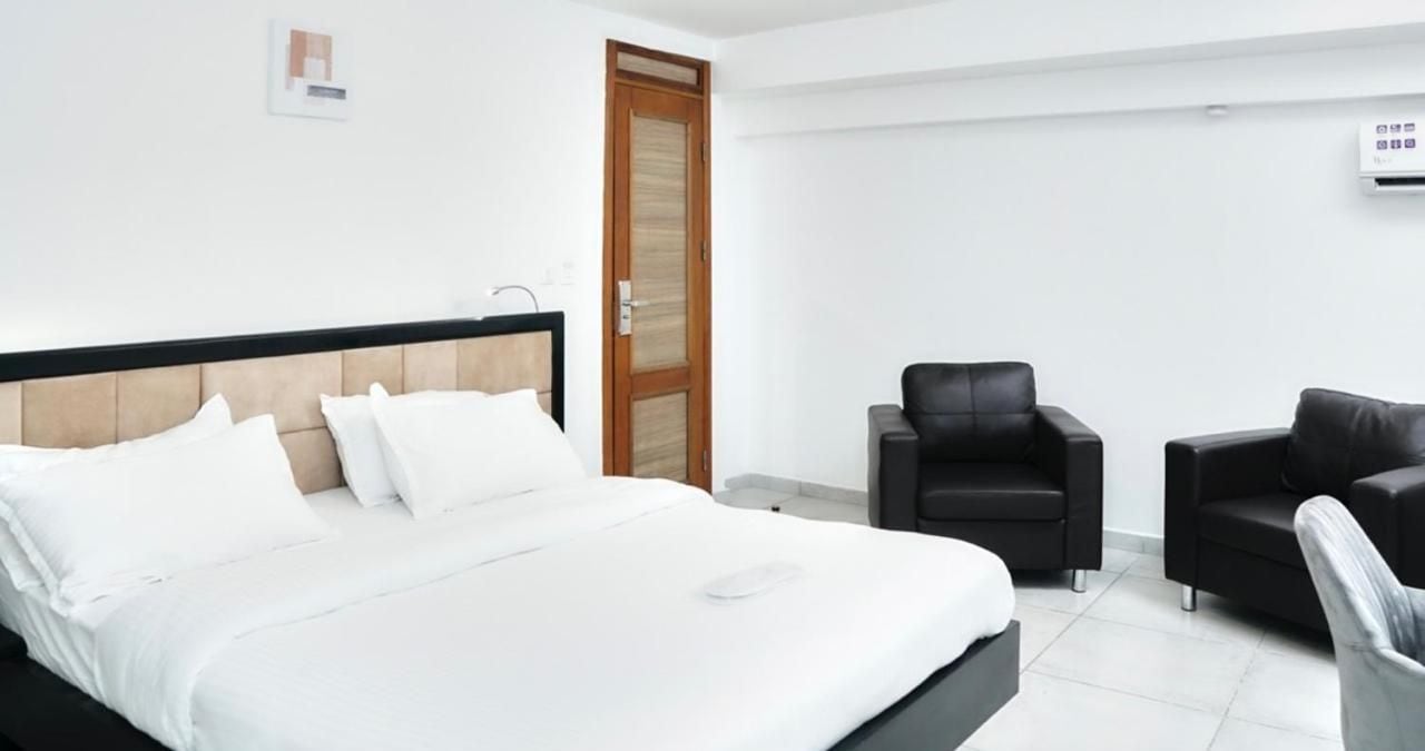 Una habitación de hotel con una cama grande, dos sillas negras, una puerta de madera y una pared blanca.