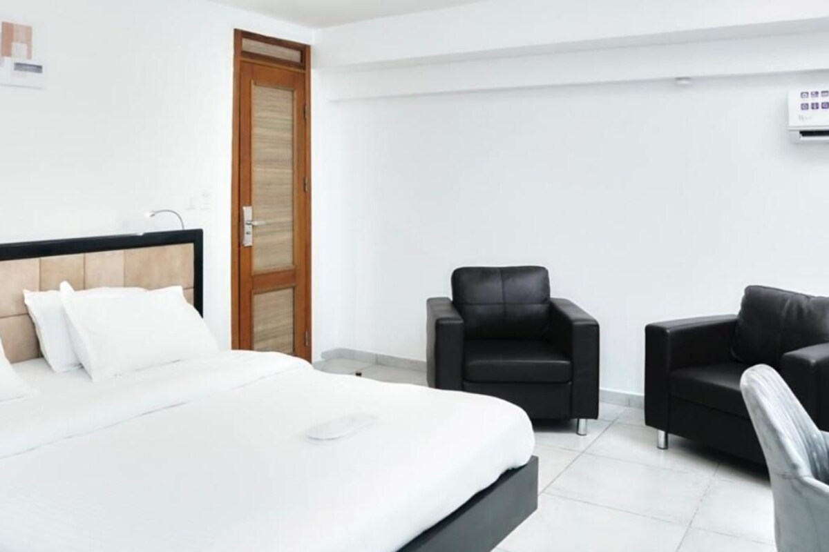 A hotel room with a large bed, two black chairs, a wooden door, and a white wall.