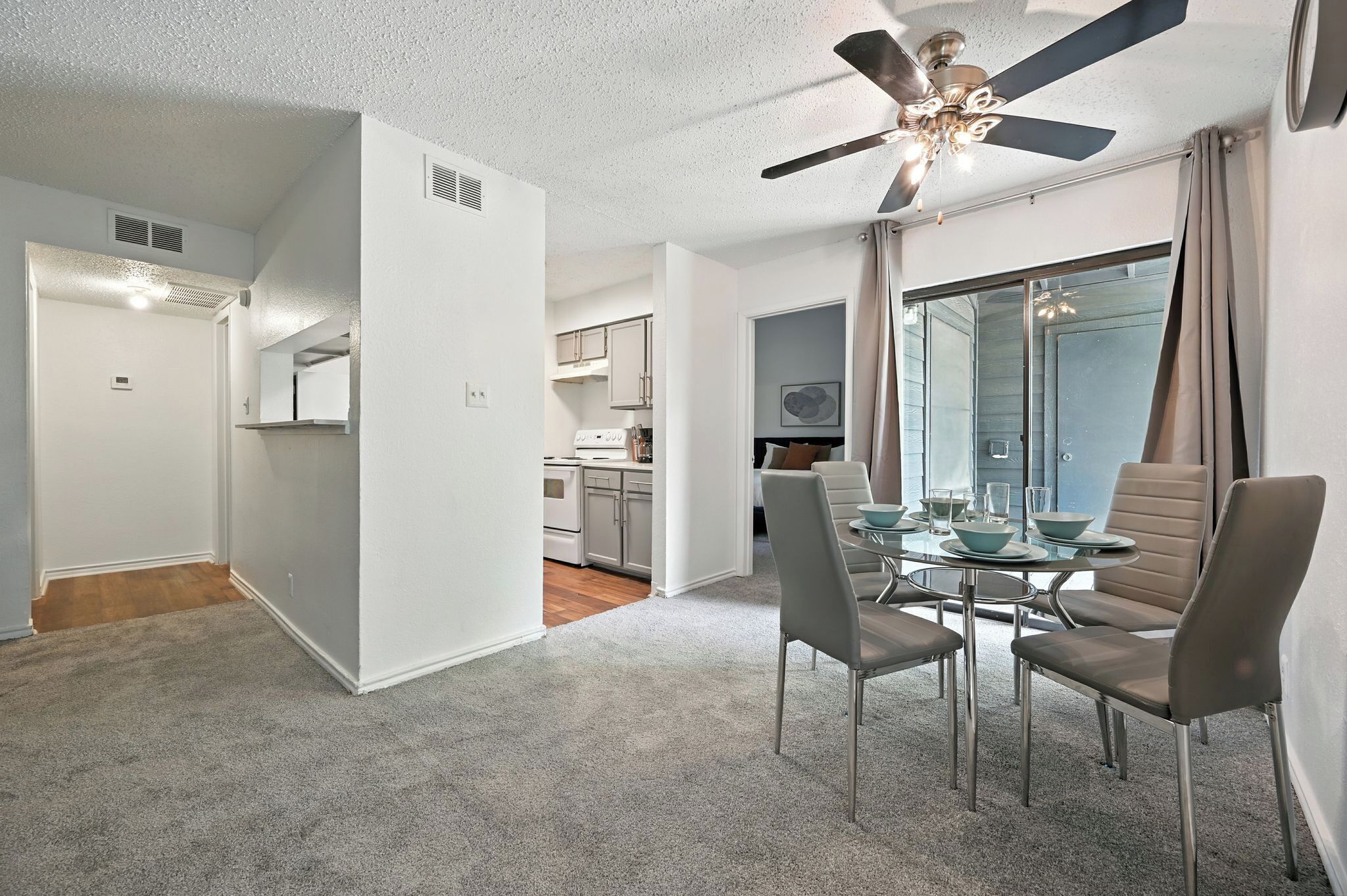 A modern apartment with a dining area featuring a round table and four chairs, a ceiling fan, and a view through a large window.