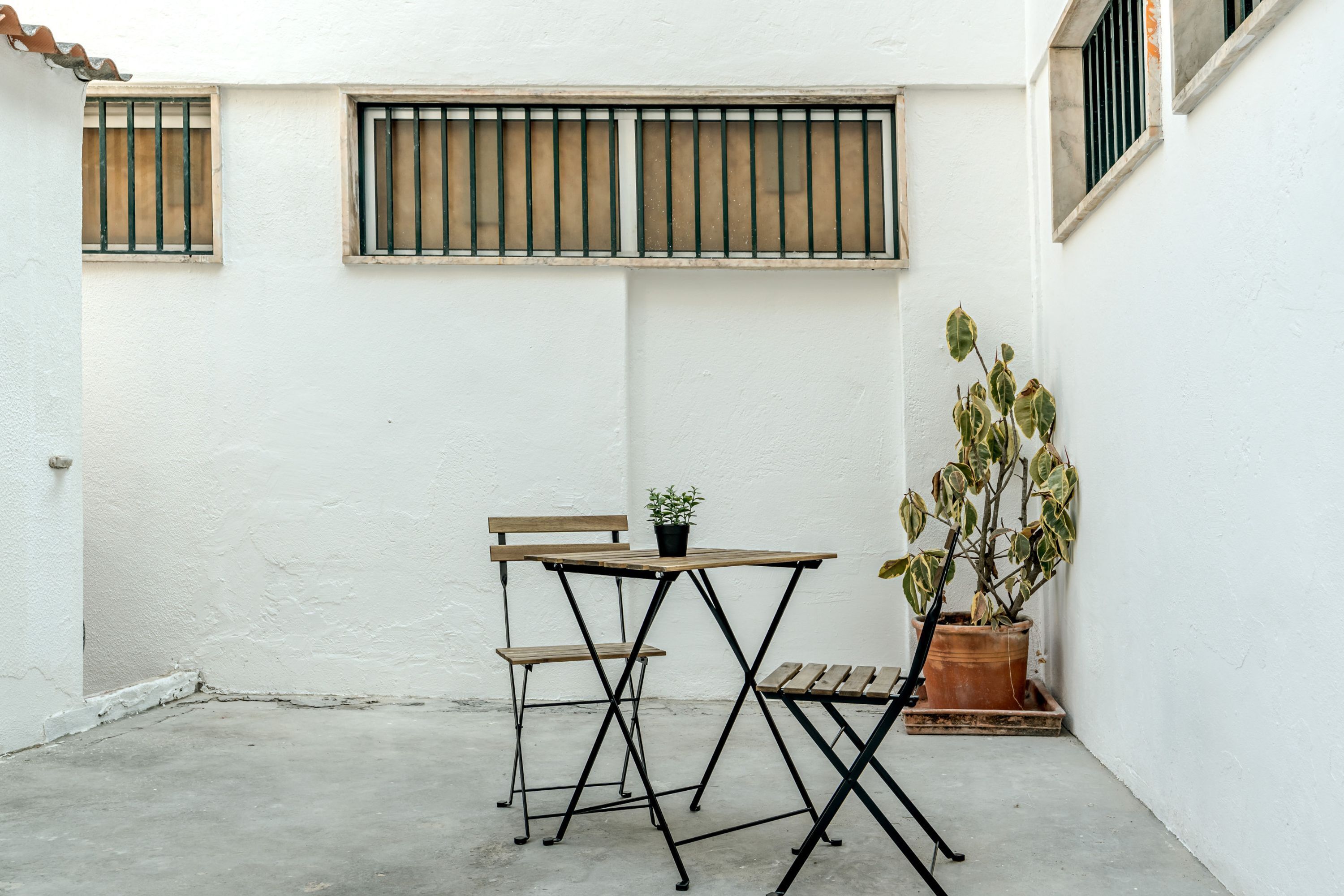 A small patio with a wooden table and chairs, a potted plant, and barred windows.