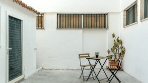 Eine kleine Terrasse mit Tisch und Stühlen, einem Blumentopf und einem vergitterten Fenster.