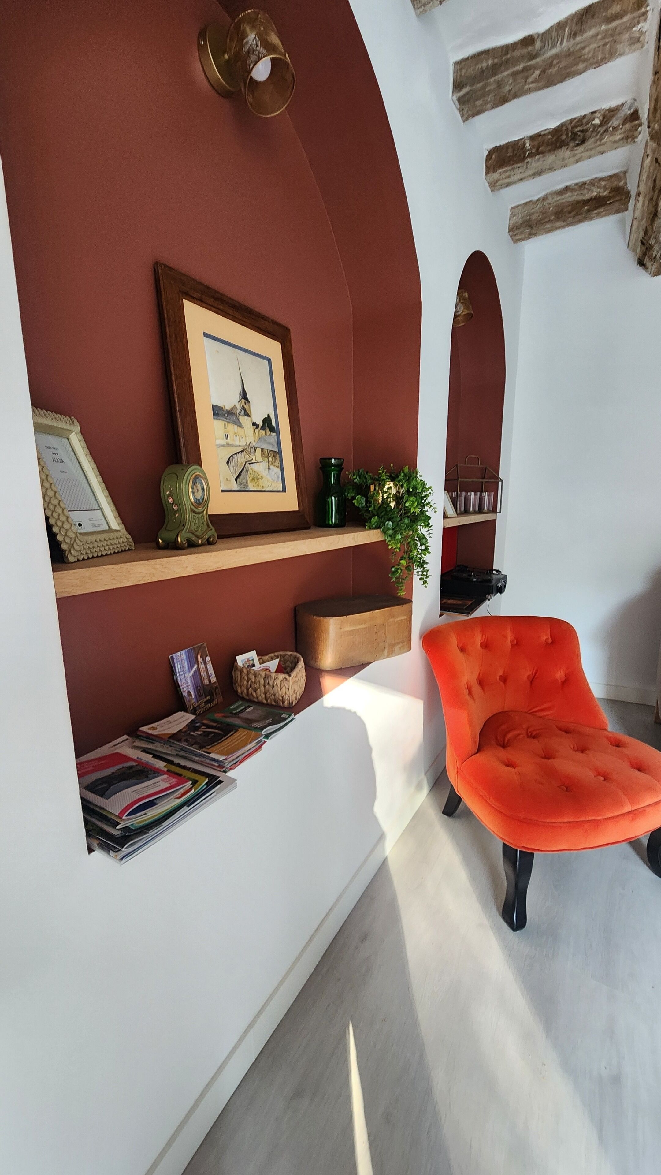 Ein Zimmer mit roter Wand, einem gerahmten Bild, einem Bücherregal, einer Pflanze und einem leuchtend orangefarbenen Sessel.