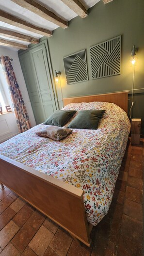 A bed with a floral bedspread, two pillows, and a wooden headboard. There are two wall-mounted light fixtures and a door in the background. - The steeple house in the heart of Mayenne. Peace and quiet (Chémeré-le-Roi)