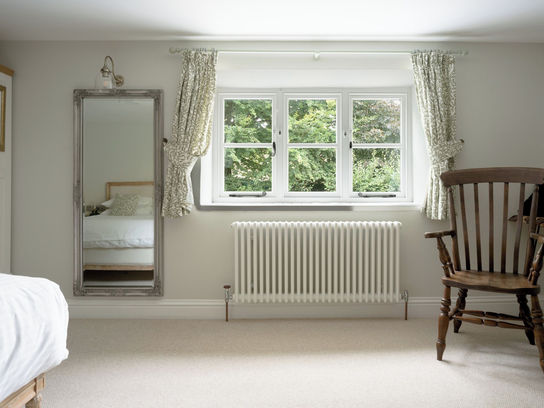 A bedroom with a large mirror, a window with curtains, a radiator, and a wooden chair.