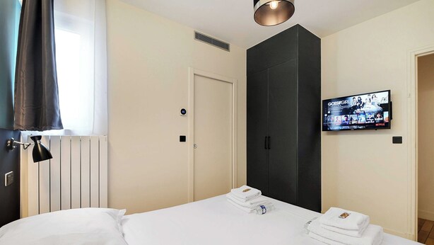 A hotel room with a bed, a TV mounted on the wall, a door, and a window with curtains. - Modern Apartment - 2br/8p - Near Opera Garnier (Paris)