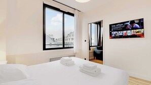A hotel room with a large window, a bed with white linens, and a wall-mounted TV displaying 'The Office'. - Modern Apartment - 2br/8p - Near Opera Garnier (Paris)