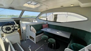 A boat's interior with a steering wheel, a green cushioned bench, and two stools. - IRIS BATEAU DOUILLET (Nevers)