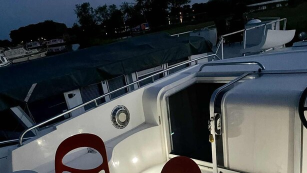 A boat deck with a table and two chairs, a storage compartment, and a view of the water and land in the background. - IRIS BATEAU DOUILLET (Nevers)