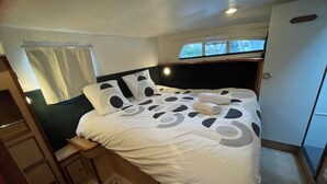 A neatly made bed with a white duvet and black circular patterns, a bedside table with a lamp, a window with curtains, and a door to the right. - IRIS BATEAU DOUILLET (Nevers)