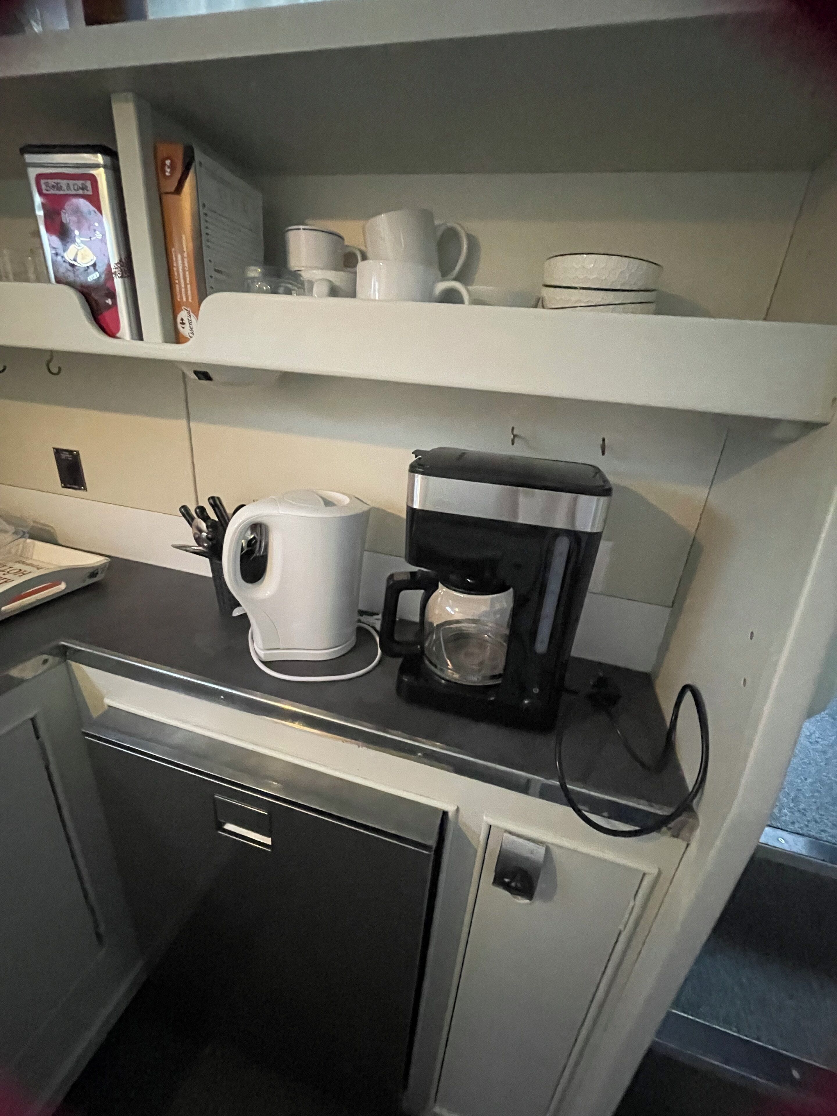 Une petite cuisine équipée d’une machine à café, d’une bouilloire et d’une étagère avec des tasses et une boîte.