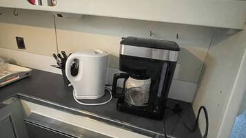 Une petite cuisine équipée d’une machine à café, d’une bouilloire et d’une étagère avec des tasses et une boîte.