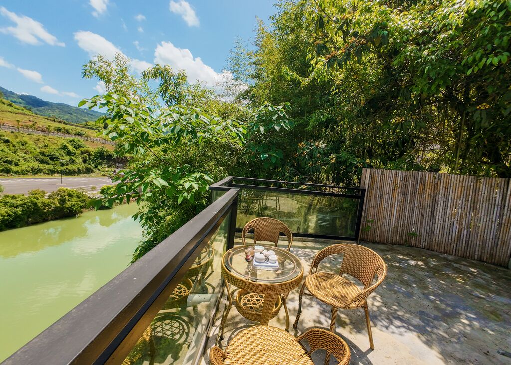 A balcony with wicker chairs and a table, overlooking a green river and lush greenery.