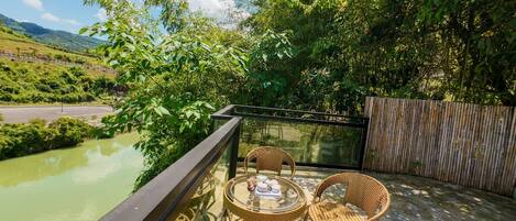 Een balkon met rieten stoelen en een tafel, met uitzicht op een groene rivier en weelderige begroeiing.