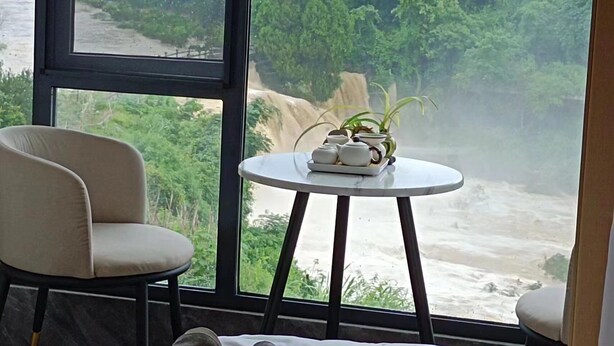 Una habitación con un ventanal grande que da a una cascada y follaje. Hay una mesa redonda con una planta y teteras encima.