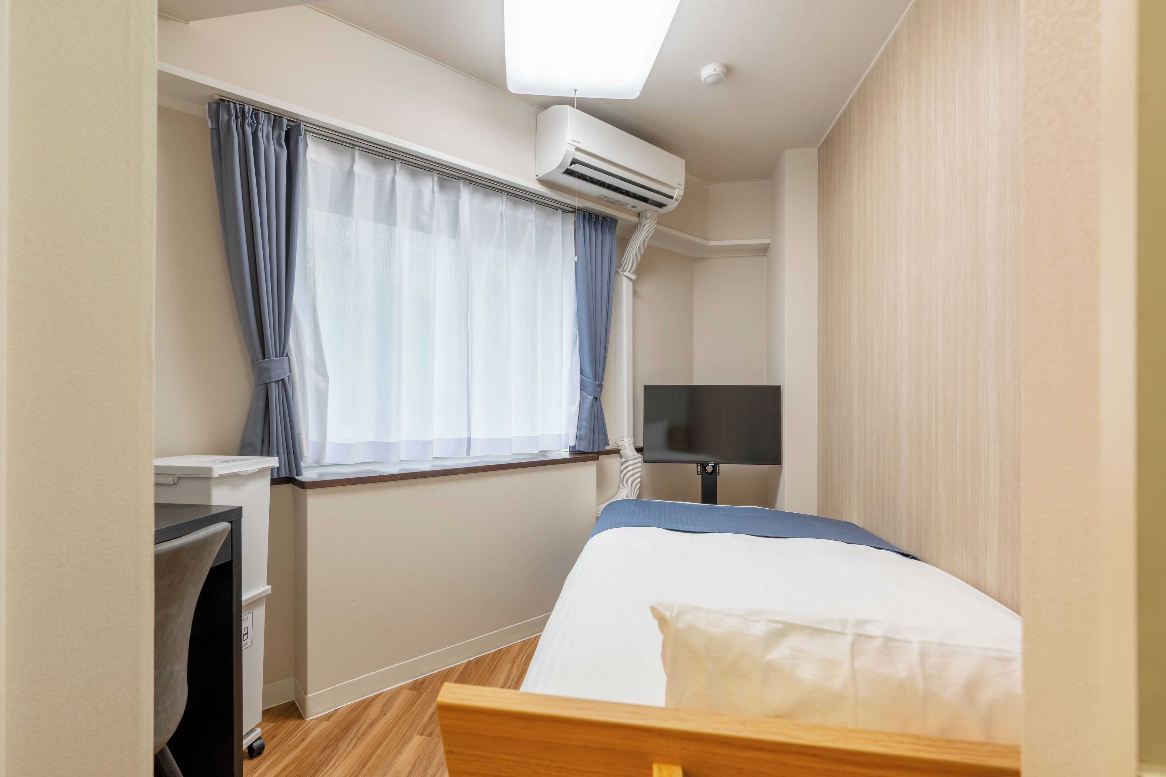 A compact hotel room with a bed, a desk, an air conditioning unit, a television, and a window with curtains.