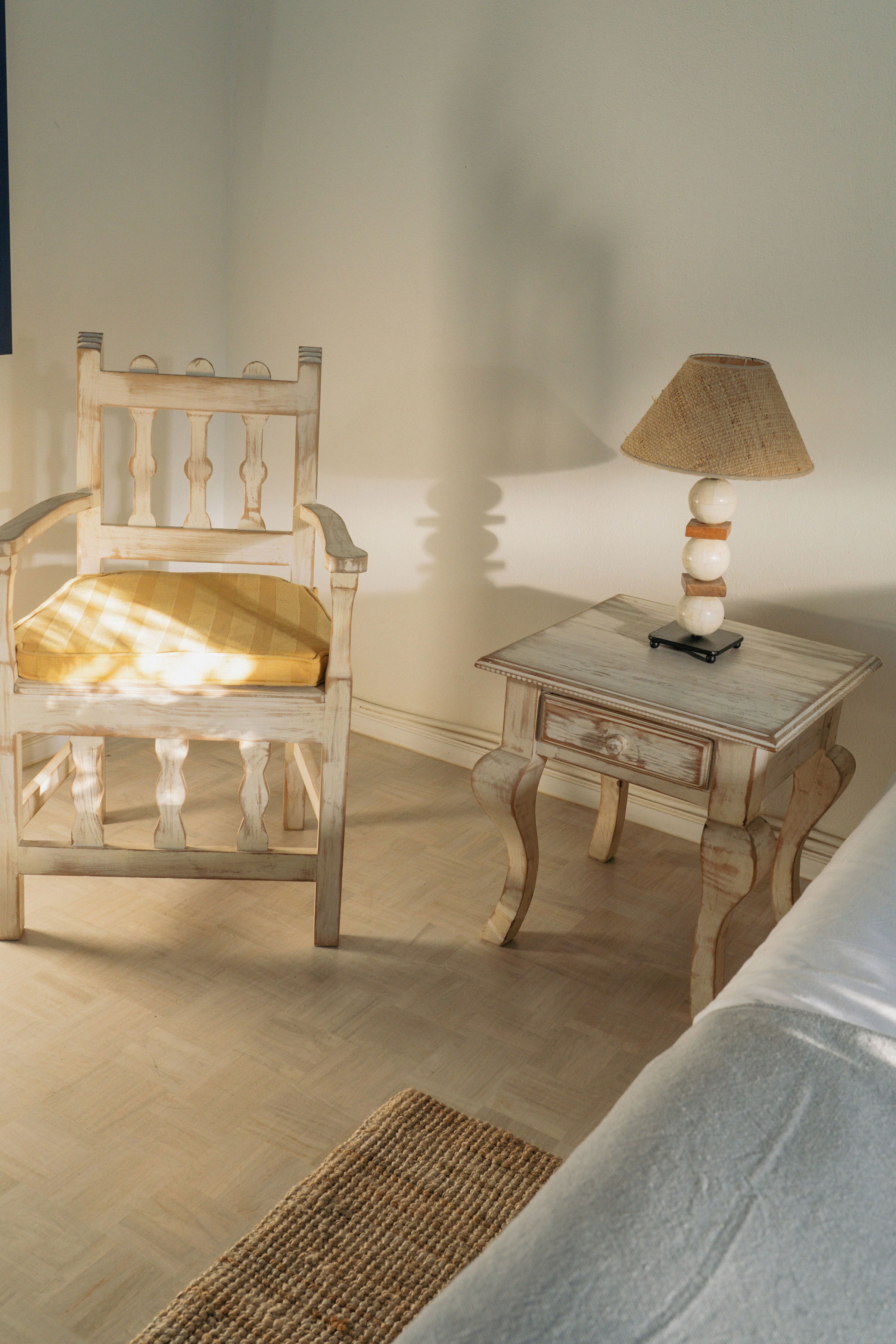 Quarto com cadeira de madeira e almofada, mesa pequena com abajur, e uma cama.