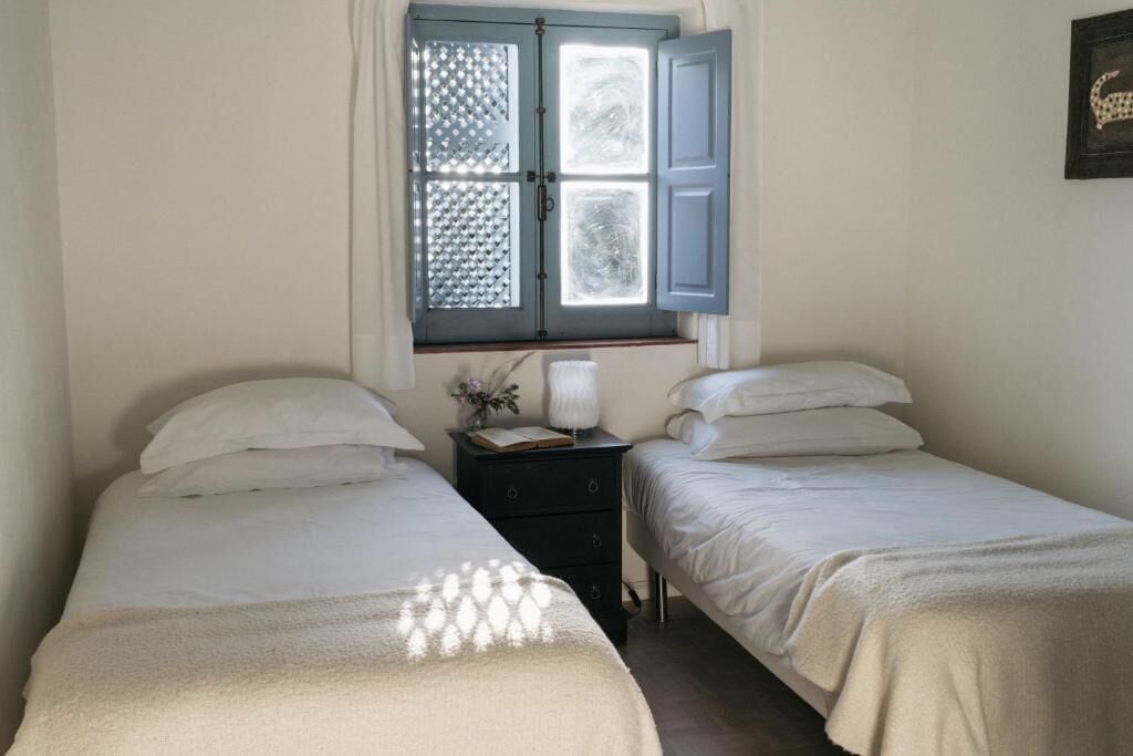 A bedroom with two single beds, a nightstand, a window with blue shutters, and a wall-mounted picture.