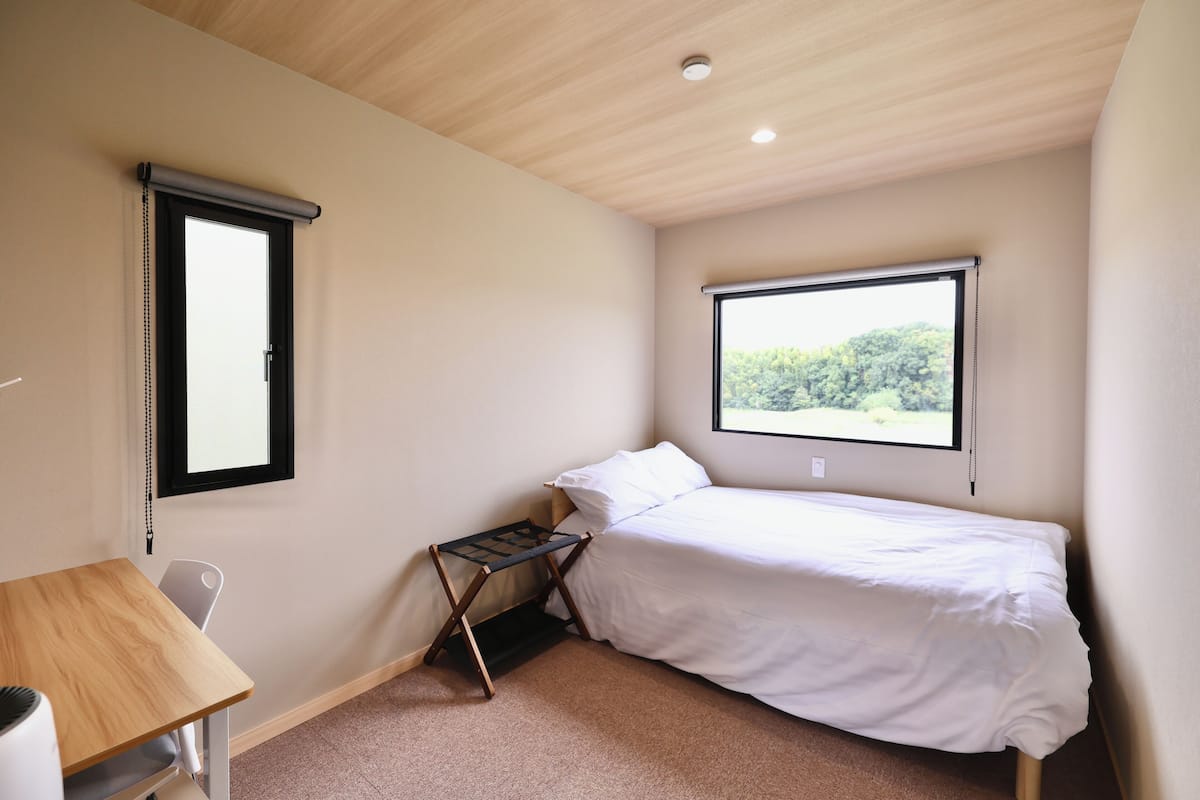 Quarto com uma cama de solteiro, uma escrivaninha e janela com vista para árvores.