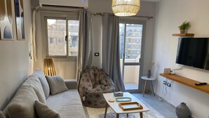 A modern living room with a sofa, a patterned armchair, a coffee table, and a television on a wooden shelf. - Sunny Haven 1BR Apartment: A Luxurious Retreat near Cairo Airport (Cairo Governorate)
