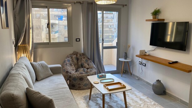 A modern living room with a sofa, a bean bag, a coffee table, a TV, and a balcony with a view of buildings. - Sunny Haven 1BR Apartment: A Luxurious Retreat near Cairo Airport (Cairo Governorate)