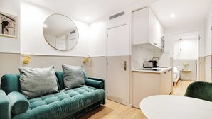 Ett modernt rum med en turkosfärgad soffa, en rund spegel, en liten köksdel och ett matbord.