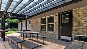 An outdoor patio with a stone wall, a black door, and a covered area with a table and benches. - Elevated Austin Escape | 4BR + Pool (Austin)