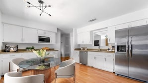 A modern kitchen with white cabinets, a stainless steel refrigerator, a glass dining table, and a contemporary light fixture. - Elevated Austin Escape | 4BR + Pool (Austin)