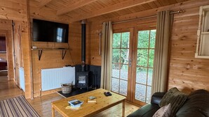 A cozy cabin interior with a wooden floor, a sofa, a coffee table with books, a TV mounted on the wall, and a wood-burning stove.
