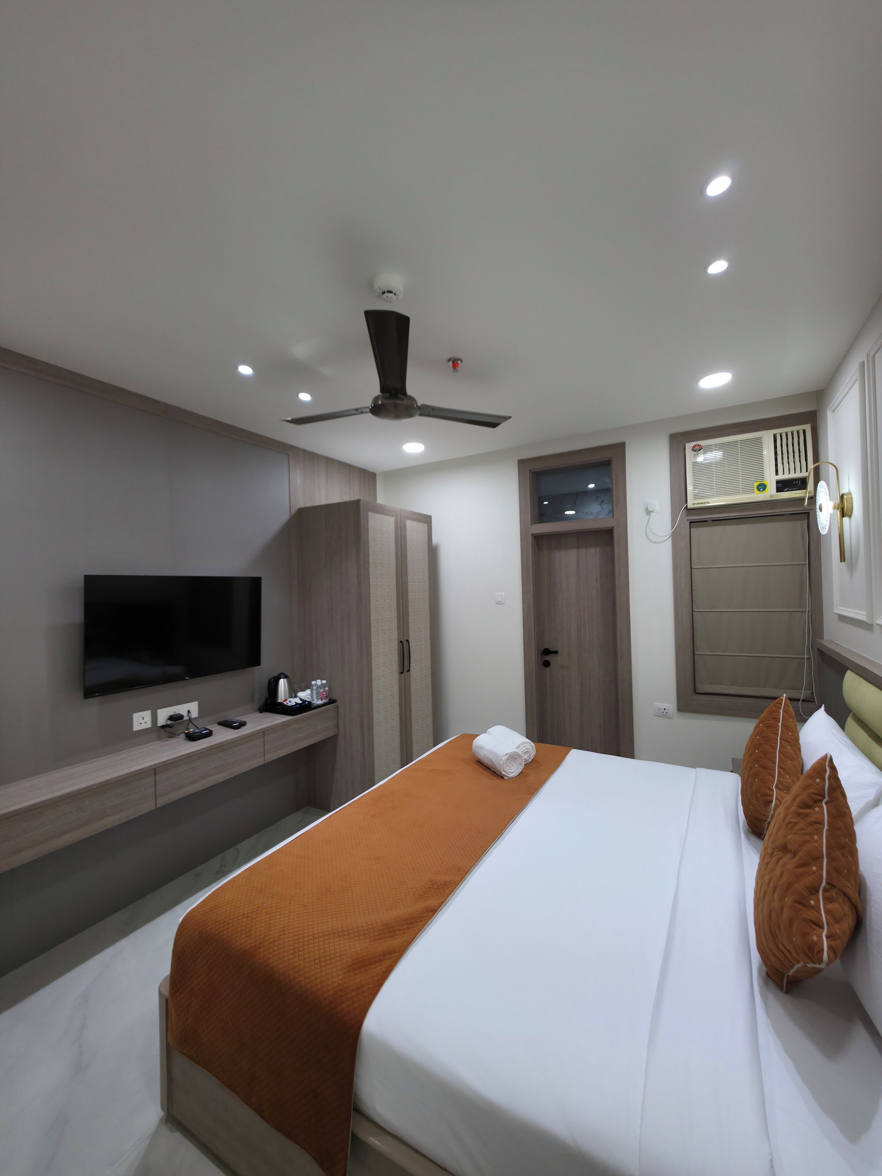 Una habitación de hotel moderna con cama, televisor de pantalla plana, ventilador de techo y ventana con unidad de aire acondicionado.