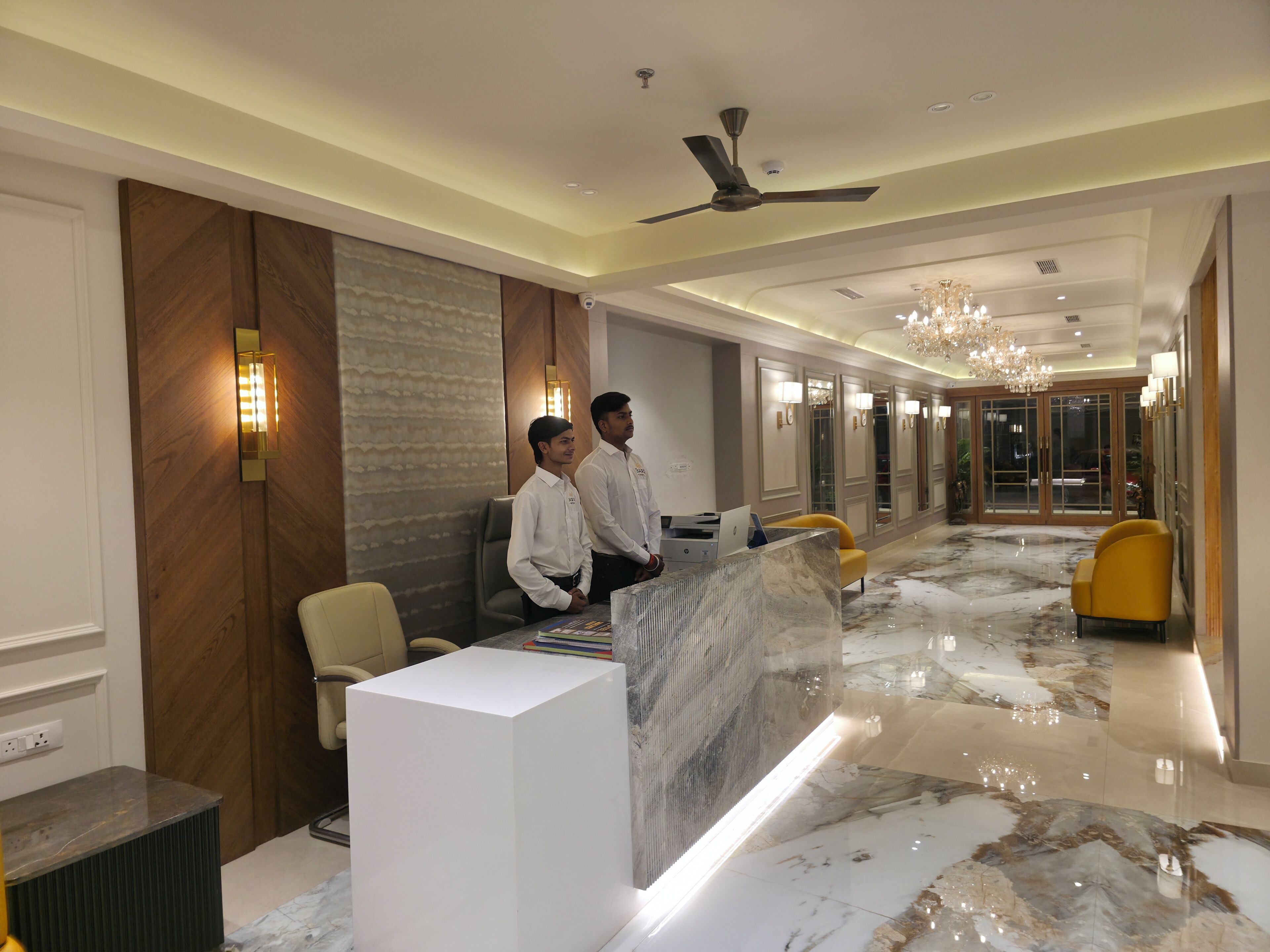A hotel reception area with a marble floor, a reception desk, a chandelier, and a ceiling fan.