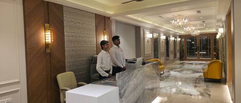 A hotel reception area with a marble floor, a reception desk, a chandelier, and a ceiling fan.