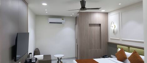 A modern hotel room with a bed, bedside table, TV, air conditioning unit, ceiling fan, and a small round table.