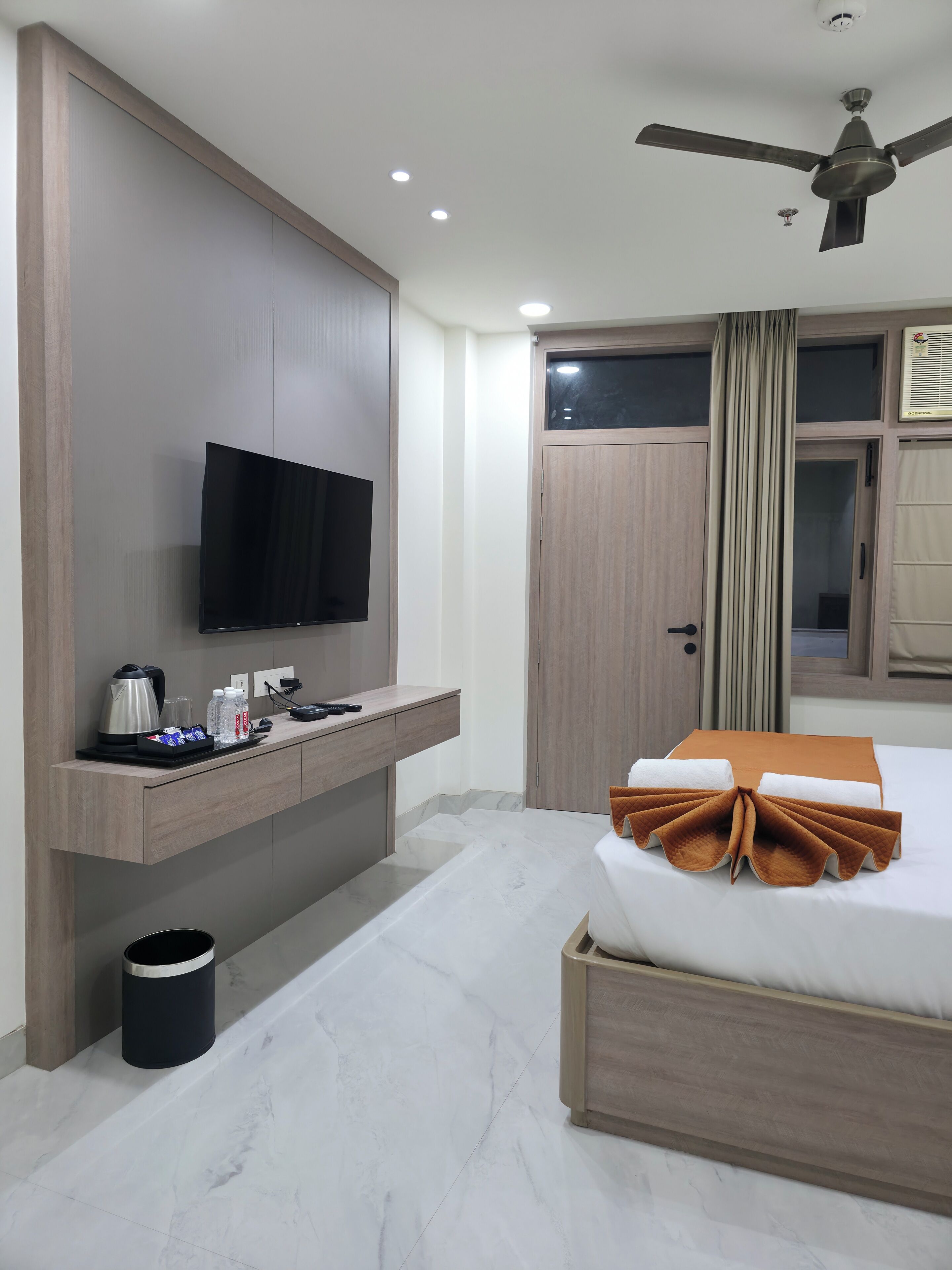 A hotel room with a bed, a TV mounted on the wall, a bedside table with a kettle and water bottles, a ceiling fan, and a window with curtains.