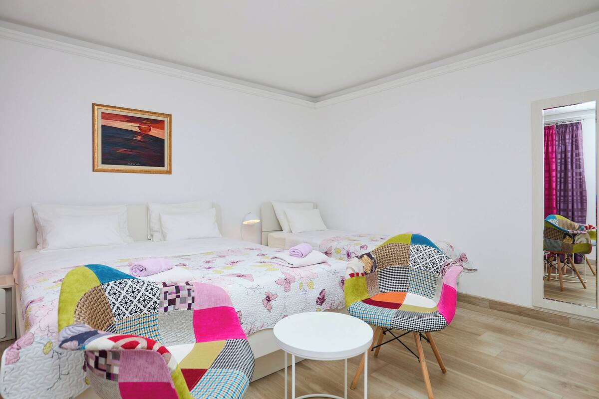 A bedroom with a bed, colorful chairs, a small table, and a painting on the wall.