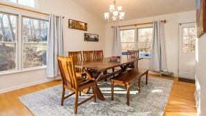 Une salle à manger avec une grande table en bois, des chaises et un lustre. Il y a deux fenêtres avec des rideaux, un tapis sur le sol et une porte avec un paillasson.