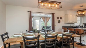 Un comedor con una mesa de madera dispuesta para seis personas, rodeada de sillas oscuras. Se observa una zona de cocina con armarios blancos y una encimera a la derecha.