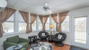Una sala de estar acogedora con un sofá verde, dos sillones, una mesa de centro y un tapete redondo. Hay tres ventanas con cortinas color beige, un ventilador de techo y una lámpara sobre una mesita.