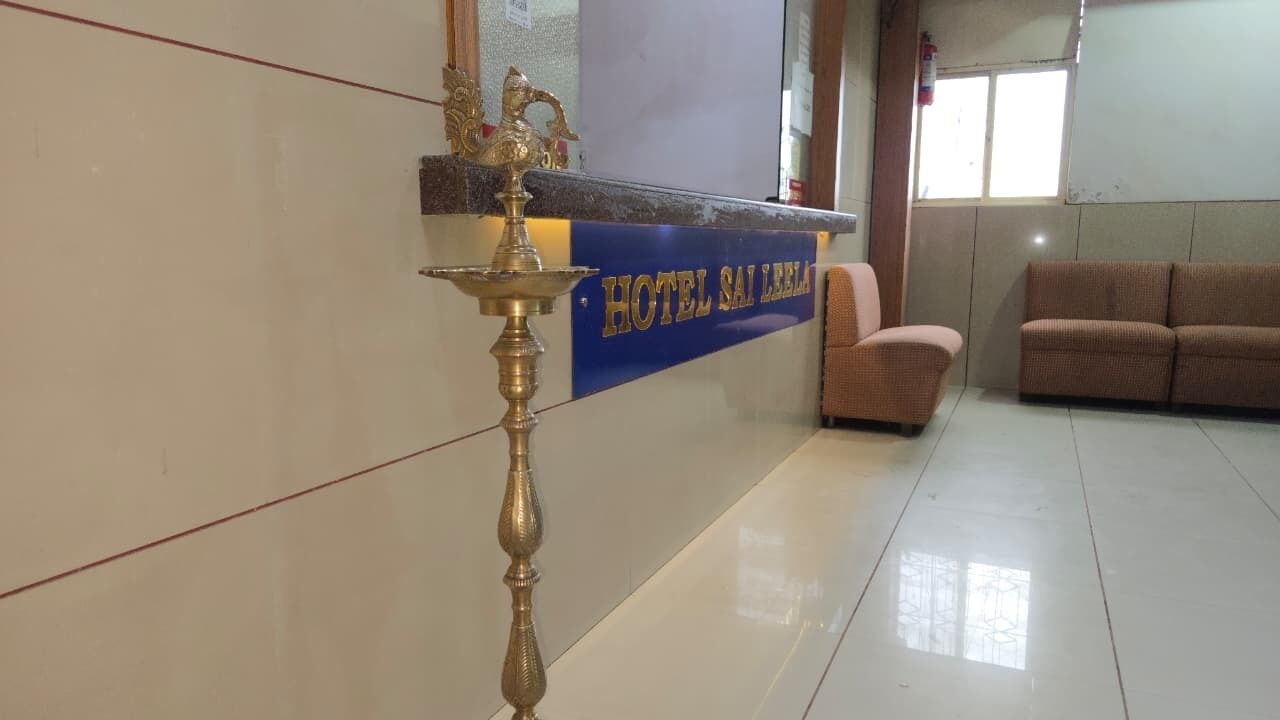 A reception area with a counter labeled "HOTEL SAI LEELA," a traditional brass lamp, a pink chair, and a brown sofa.