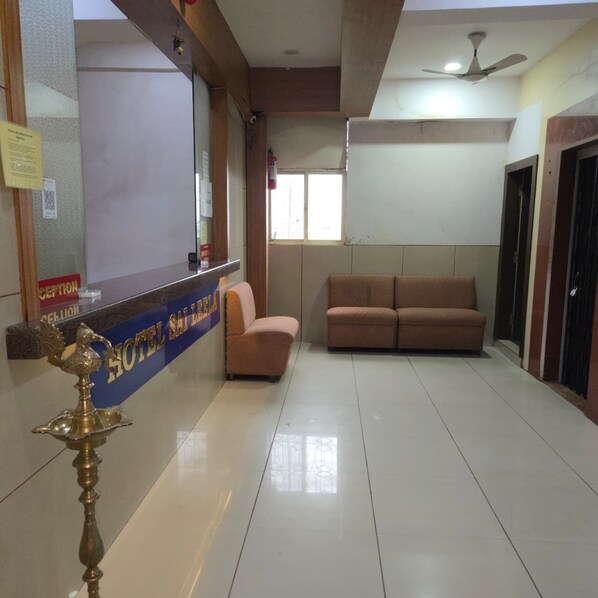 A hotel reception area with a reception desk, two sofas, a ceiling fan, and a decorative brass lamp. - Hotel Sai Leela (Ambernath)