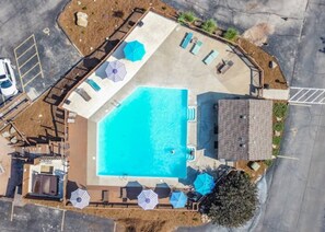 An aerial view of a swimming pool area with a rectangular pool, lounge chairs, umbrellas, and a small building.