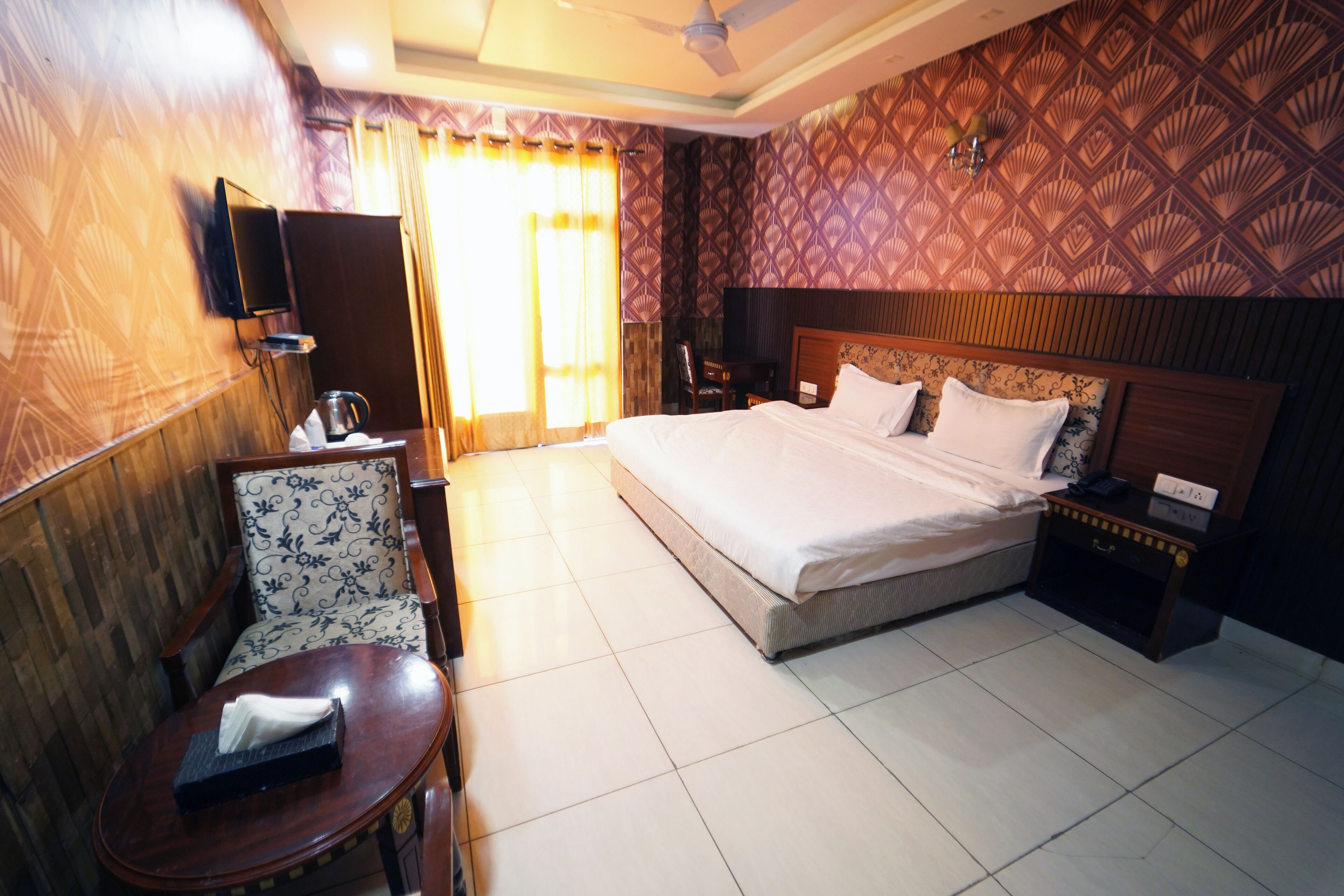 A hotel room with a large bed, a wooden headboard, a bedside table with a lamp, a chair, and a TV mounted on the wall.