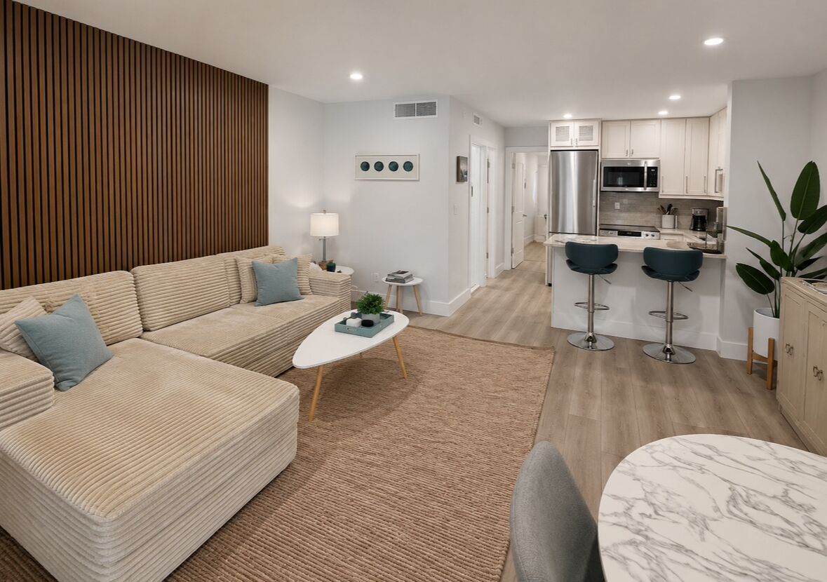 A modern living room with a sofa, coffee table, and a small kitchen area with a refrigerator, microwave, and bar stools.