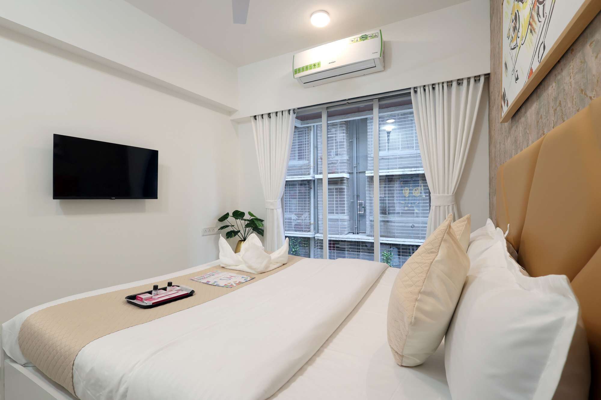 A hotel room with a bed, a flat-screen TV, an air conditioner, a window with curtains, and a potted plant.